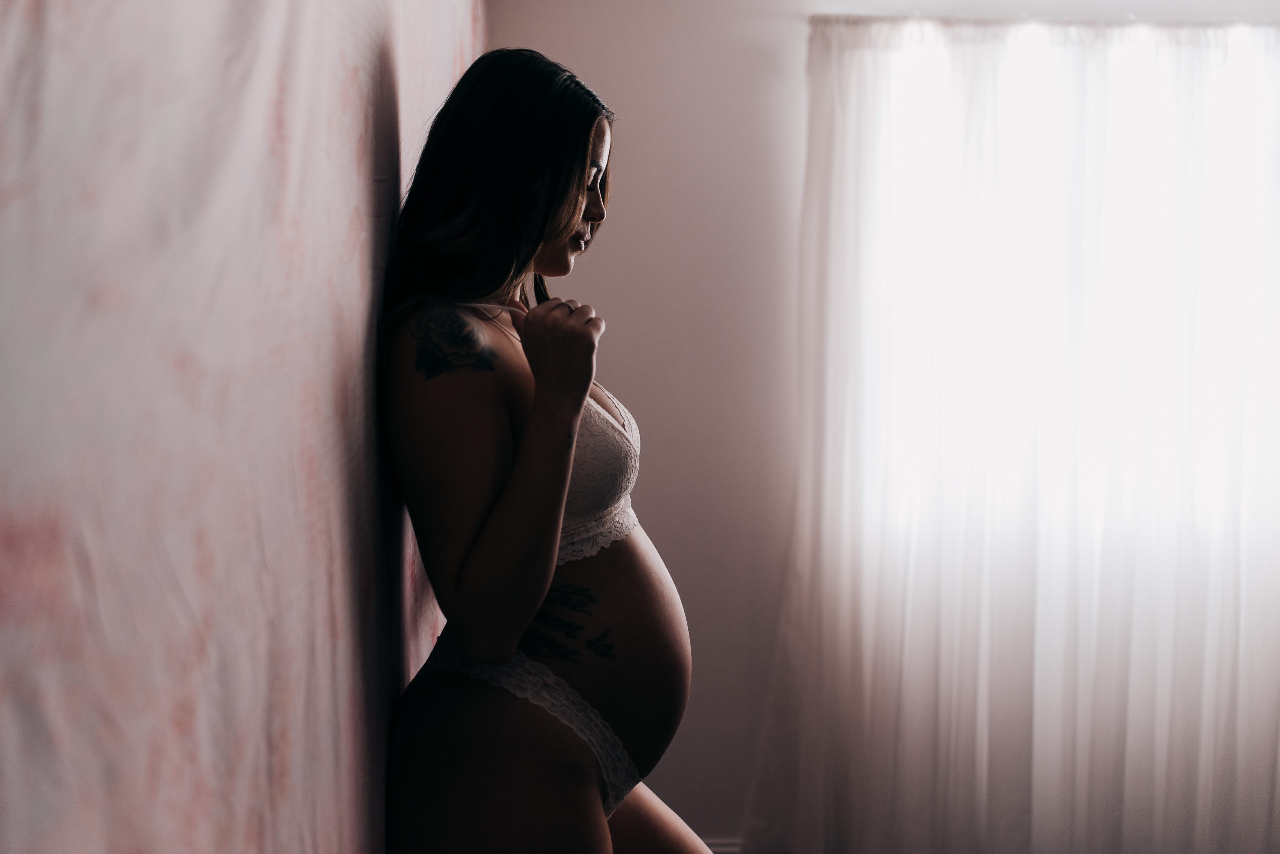 pregnant woman during her boudoir session wearing a bra and panty set while leaning against a floral pink wall