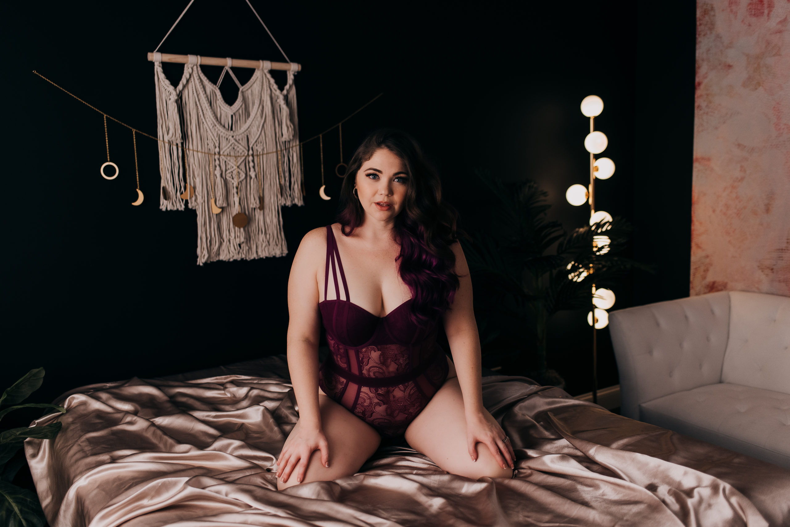 woman wearing a deep purple bodysuit while posing on a bed with champagne colored sheets