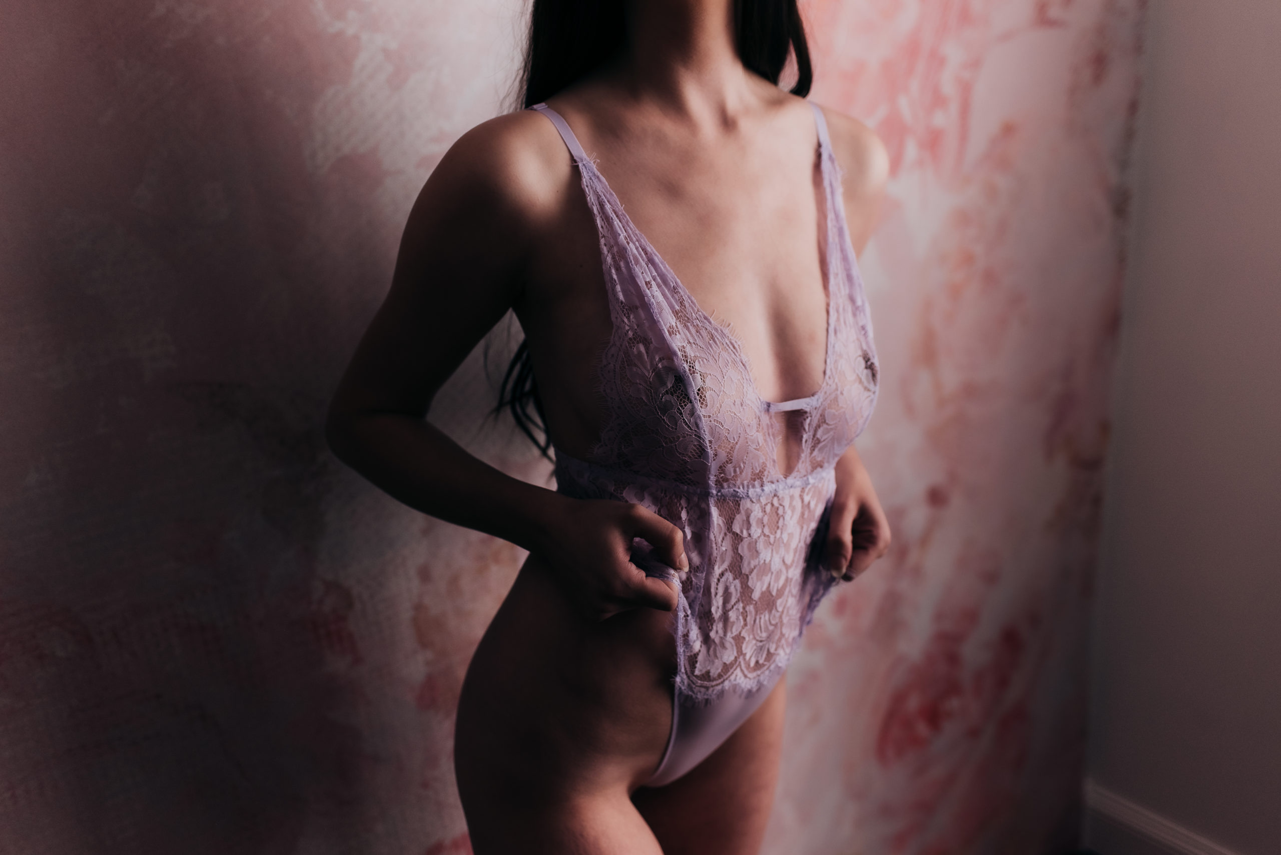 woman during her boudoir session wearing a lilac teddy and standing against a pink floral wall