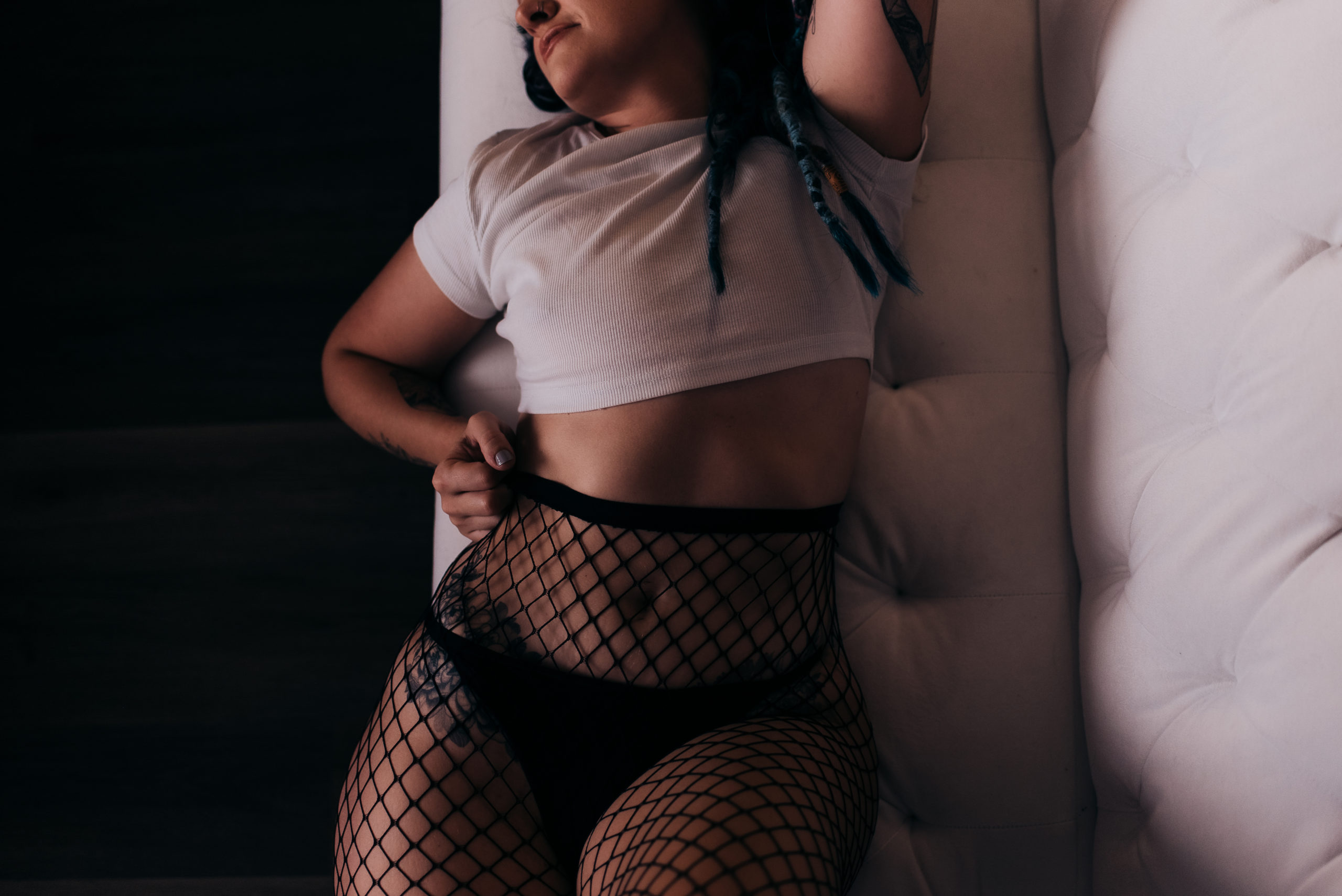 woman during her boudoir session wearing a white crop top, black underwear, and a pair of fishnets while laying on a white couch